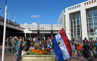 Foto der Versammlung zum 420 Smoke-Out vor Stadthalle und Rathaus in Amsterdam. Im Vordergrund eine niederlndische Flagge mit Anonymous-Logo